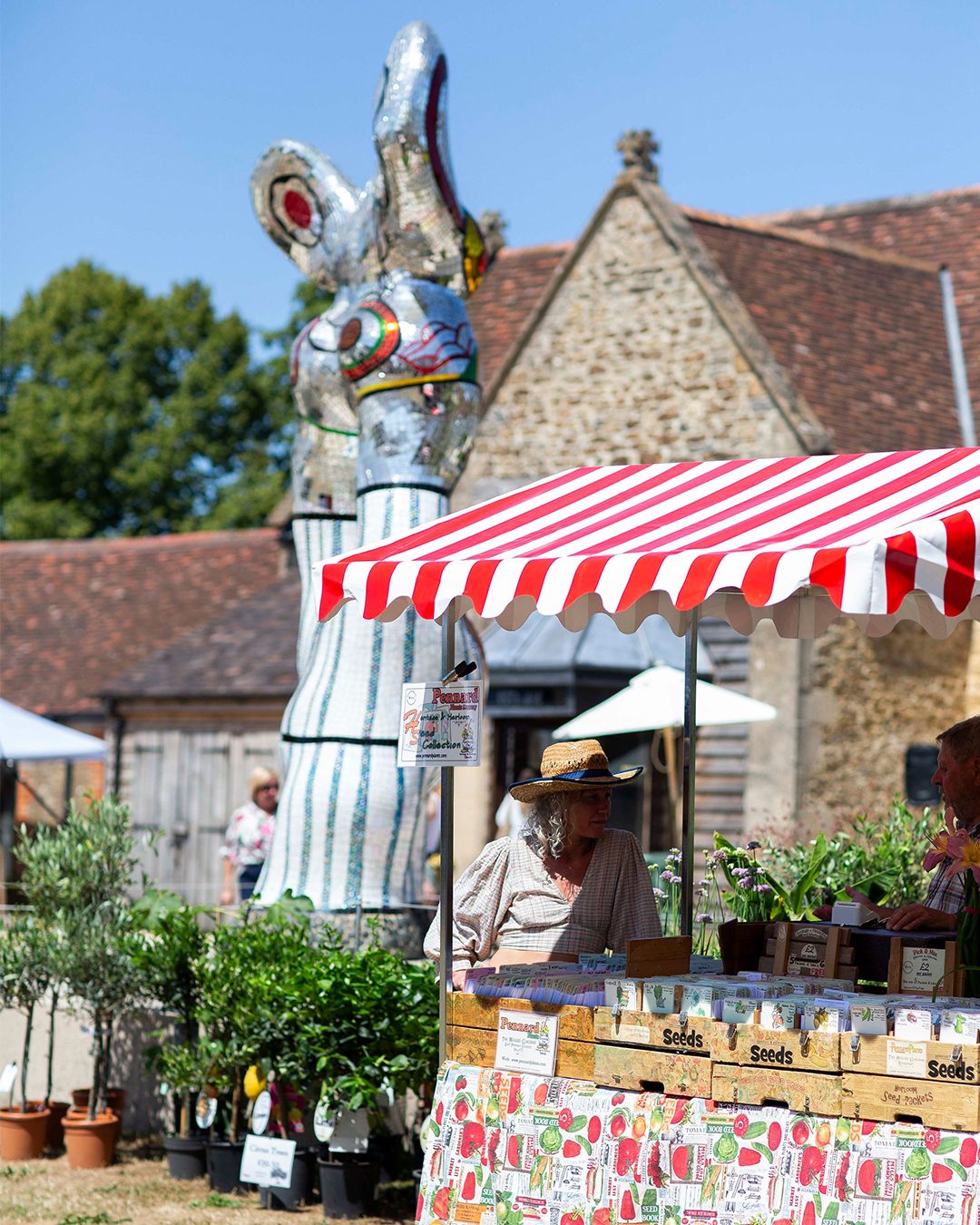 A striped market stall and plants at the Hauser & Wirth Somerset summer fête, beneath a tall mirror-mosaic sculpture